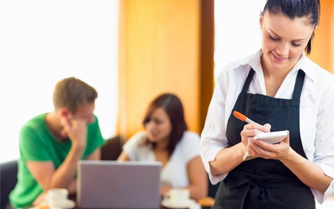 A person in an apron writes down an order with two people on laptops sitting in the background