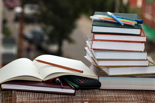 A stack of textbooks topped by various pens, beside a closed leather notebook topped by an open graph paper notebook with a pencil on top, various pens and a calculator under the open notebook