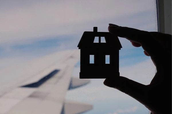 A silhouette of a small house model is held against the backdrop of an airplane wing and sky visible through a window.