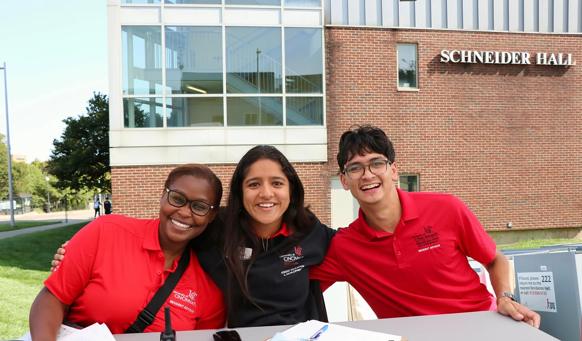 Three RAs in red University of Cincinnati shirts are smiling in front of Schneider Hall.