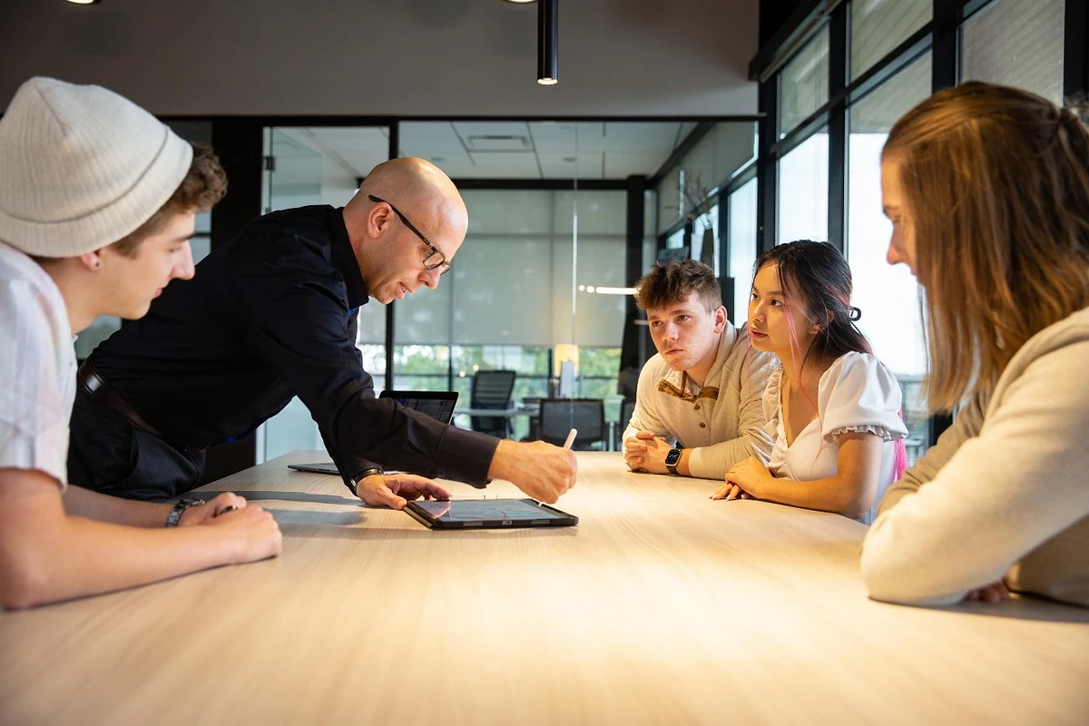 A group of people gathers around a table as one person leans over to use a tablet