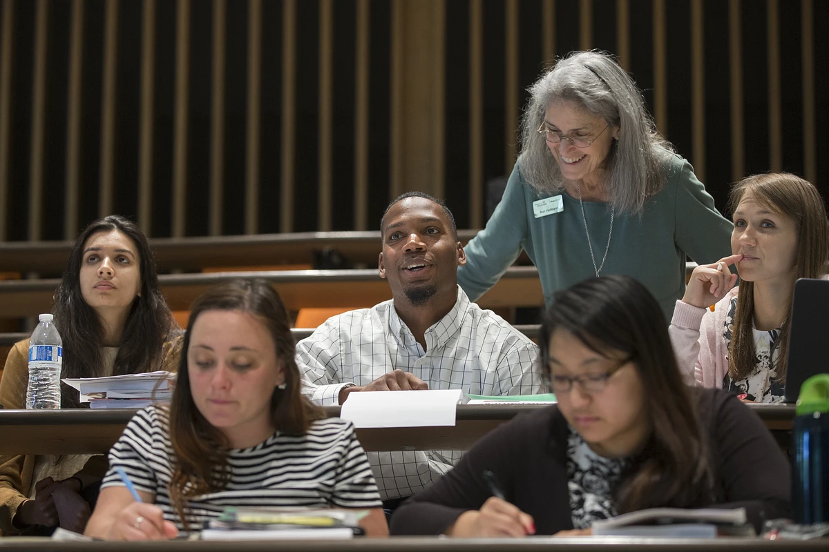 As faculty member looks on as students seated in the classroom have a discussion